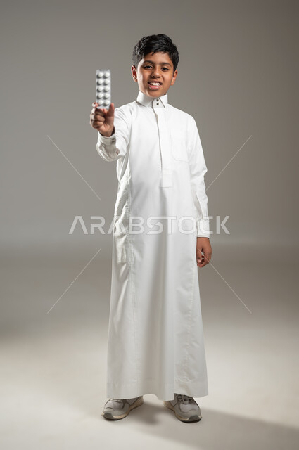 الإصابة بالمرض والتوعك, النظر للكاميرا بإيماءات السرور, مفهوم الصحة والأمراض والعلاجات الطبية, بروتريه لفتى عربي خليجي سعودي يرتدي ثوبه الأبيض التقليدي يمسك بيده برشام الدواء, جسم بالطول الكامل, خلفية رمادية, محتوى حساس, استخدام حساس