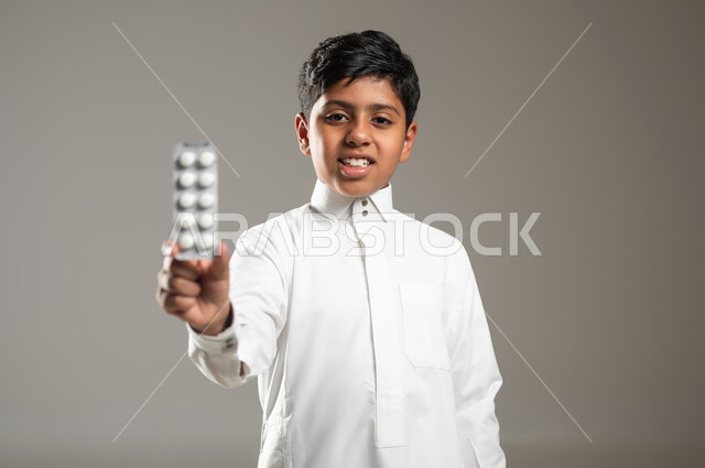 النظر للكاميرا بإيماءات السرور, مفهوم الصحة والأمراض والعلاجات الطبية, بروتريه لفتى عربي خليجي سعودي يرتدي ثوبه الأبيض التقليدي يمسك بيده برشام الدواء, الإصابة بالمرض والتوعك, خلفية رمادية, محتوى حساس, استخدام حساس