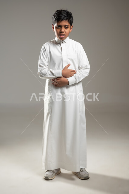 وضع اليدين على البطن و النظر للكاميرا بإيماءات تدل على الألم والامتعاض, بروتريه لفتى عربي خليجي سعودي يرتدي ثوبه الأبيض التقليدي يحتاج إلى العلاج والراحة, خلفية رمادية, جسم بالطول الكامل, محتوى حساس, استخدام حساس