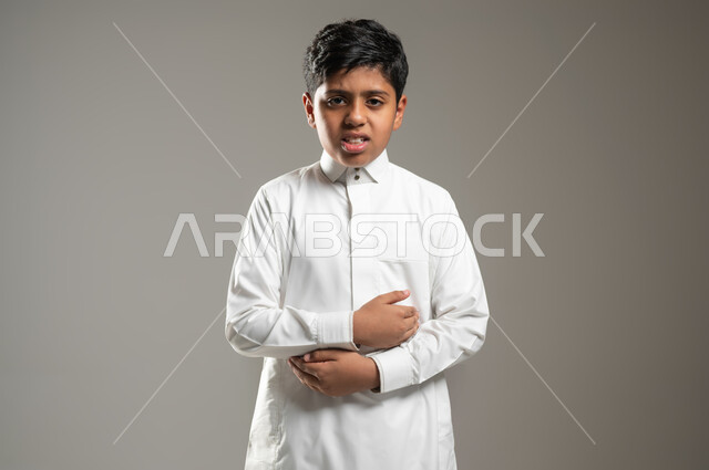 الإصابة بوعكة صحية, النظر للكاميرا بإيماءات تدل على وجود الآلام والأوجاع, بروتريه لفتى عربي خليجي سعودي يرتدي ثوبه الأبيض التقليدي يضع يديه على بطنه بتعبيرات الامتعاض وعدم الراحة, خلفية رمادية, محتوى حساس, استخدام حساس