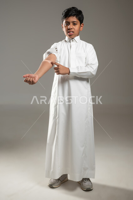 The role of medical bandages in relieving pain, looking at the camera with gestures indicating the presence of pain, portrait of a Saudi Gulf Arab boy wearing his traditional white dress pointing with his little finger at a small sticker on his hand, gray background, full-length body, sensitive content, sensitive use