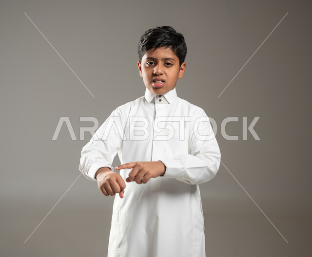 الإصابة بجروح أو حروق او التعرض لرضوض في اليد, النظر للكاميرا بإيماءات تدل على وجود الآلام والأوجاع, بورتريه لفتى عربي خليجي سعودي يرتدي ثوبه الأبيض التقليدي يشير باصبعه إلى مكان الإصابة في يده الصغيرة بتعبير الحزن, خلفية رمادية, محتوى حساس, استخدام حساس