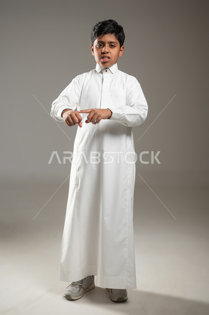 الإصابة بوعكة صحية, النظر للكاميرا بإيماءات تدل على وجود الآلام والأوجاع, بروتريه لفتى عربي خليجي سعودي يرتدي ثوبه الأبيض التقليدي يشير بأصبعه إلى مكان الإصابة في يده الصغيرة بتعبير الحزن, خلفية رمادية, جسم بالطول الكامل, محتوى حساس, استخدام حساس