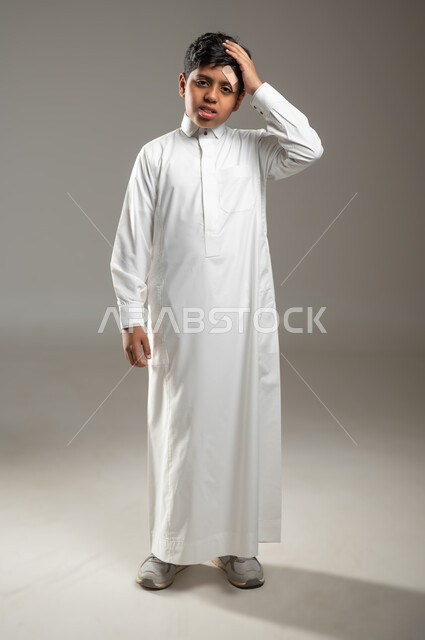 النظر إلى الكاميرا بإيماءات تدل على وجود الآلام والأوجاع, بورتريه لفتى عربي خليجي سعودي يرتدي ثوبه الأبيض التقليدي يضع يده على رأسه تعبيراً عن الألم والتعب الذي حصل له, خلفية رمادية, جسم بالطول الكامل, محتوى حساس, استخدام حساس