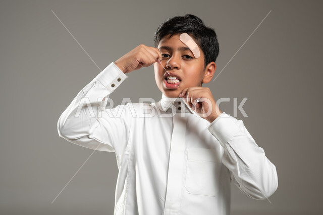 الشعور بالألم والأوجاع بعد التعرض لإصابات وجروح في الرأس والوجه, بورتريه لولد عربي خليجي سعودي يلبس الثوب البيض التقليدي ويضع اللصاقة الطبية على جبينه, الإشارة إلى مكان الإصابة في وجهه بإيماءات حزن وامتعاض, خلفية رمادية,  محتوى حساس, استخدام حساس