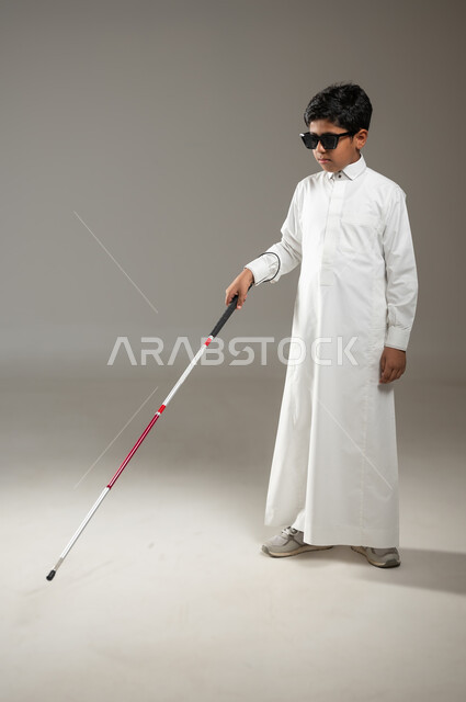العصا البيضاء ودورها في توجيه المكفوف بشكل صحيح, فقدان الرؤية والبصر, مفهوم الصحة والمرض, بورتريه لولد عربي خليجي سعودي ضرير أُصيب بالعمى يرتدي النظارة الشمسية السوداء و بيده العكاز، جسم بالطول الكامل، خلفية رمادية, محتوى حساس، استخدام حساس