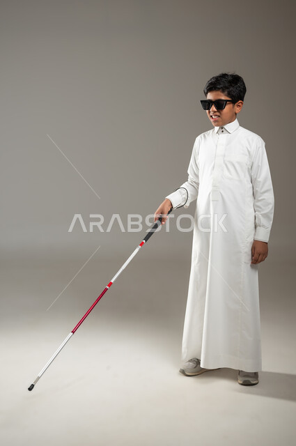 فقدان الرؤية والبصر, اعتماد الشخص الضرير على العصا البيضاء بشكل كامل في جميع تحركاته, مفهوم الصحة والمرض, بورتريه لولد عربي خليجي سعودي كفيف مصاب بالعمى ، يرتدي النظارة الشمسية و بيده العكاز، جسم بالطول الكامل، خلفية رمادية, محتوى حساس، استخدام حساس