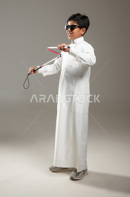 العصا البيضاء ودورها في مساعدة المكفوفين, مفهوم الصحة والمرض, بورتريه لولد عربي خليجي سعودي كفيف مصاب بالعمى، يرتدي النظارة الشمسية و بيده العكاز ، فقدان الإدراك البصري، حالة مرضية، خلفية رمادية, جسم بالطول الكامل, محتوى حساس، استخدام حساس