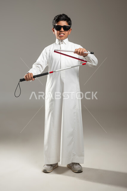 فقدان الإدراك البصري والإصابة بالعمى, مفهوم الصحة والمرض, بورتريه لولد عربي خليجي سعودي كفيف يرتدي النظارة الشمسية و بيده العكاز ( العصا البيضاء ) ، حالة مرضية ، جسم بالطول الكامل, خلفية رمادية, محتوى حساس، استخدام حساس