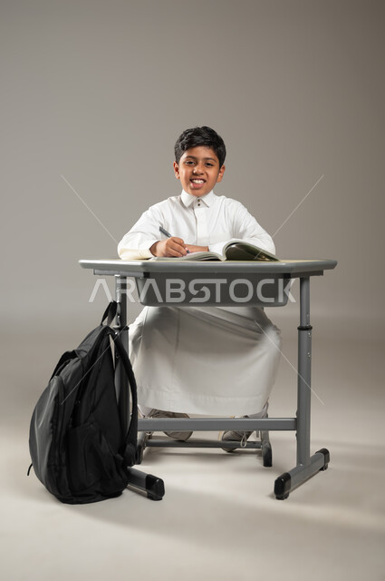 النظر إلى الكاميرا بإيماءات السعادة, الجد والإجتهاد في الدراسة والمذاكرة من أجل التفوق, بورتريه لطفل عربي خليجي سعودي يرتدي الثوب الأبيض التقليدي يجلس في مقعده ويتابع دروسه, الحقيبة المدرسية السوداء معلقة على جانب المقعد, خلفية رمادية, محتوى حساس, استخدام حساس