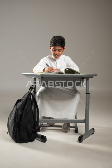متابعة المناهج الدراسية بكل مثابرة وجد من أجل النجاح, العلم والتعلم في المجتمع السعودي, بورتريه لطفل عربي خليجي سعودي يرتدي الثوب الأبيض التقليدي يجلس في مقعده ويقرأ في كتبه, الحقيبة المدرسية السوداء على جانب المقعد, خلفية رمادية, محتوى حساس, استخدام حساس