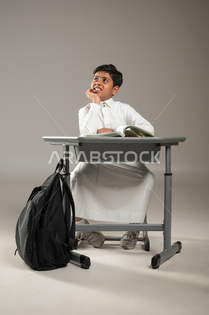 الشغف و السعي نحو النجاح والتفوق, بورتريه لطفل عربي خليجي سعودي يرتدي الثوب الأبيض التقليدي يجلس في مقعده وينظر إلى الأفق بإيماءات التفاؤل والأمل بسعادة, تحقيق الأحلام والأهداف المستقبلية, خلفية رمادية, محتوى حساس, استخدام حساس