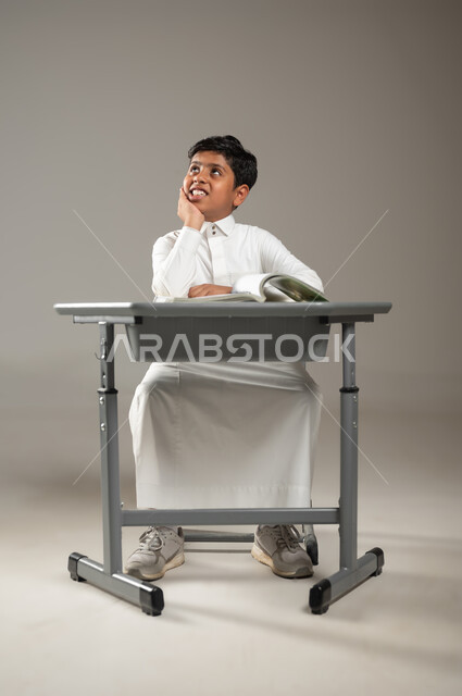 النظر إلى السماء والتحليق في عالم الأحلام المنتظرة, السعي نحو النجاح والتفوق, بورتريه لطفل عربي خليجي سعودي يرتدي الثوب الأبيض التقليدي يجلس في مقعده وينظر إلى الأفق بإيماءات السعادة,  التفكير بالخطط والأهداف المستقبلية, خلفية رمادية, محتوى حساس, استخدام حساس