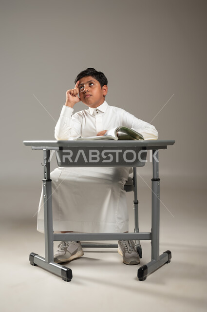 التحديق في الفراغ بإيماءات الحيرة, عدم معرفة شيء ما, , بورتريه لطفل عربي خليجي سعودي يرتدي الثوب الأبيض التقليدي يجلس في مقعده وينظر في الأفق بتمعن واضعاً يده على خده, أهمية العلم والتعلم, متابعةالمناهج الدراسية , خلفية رمادية, محتوى حساس, استخدام حساس