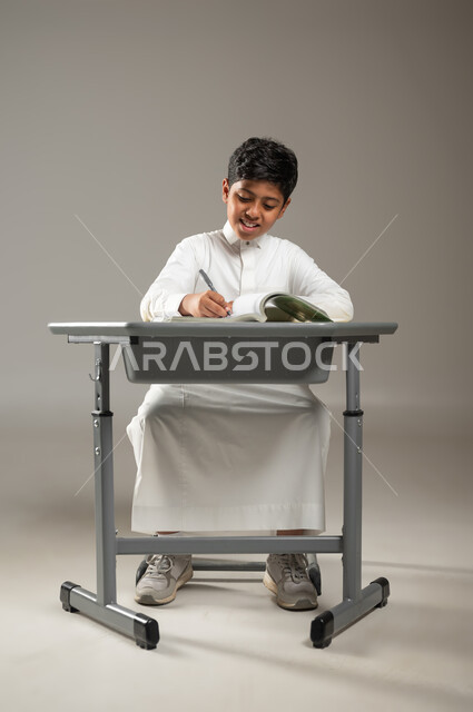 الجد والإجتهاد في الدراسة والمذاكرة من أجل التفوق, متابعةالمناهج الدراسية بكل مثابرة وعزيمة, بورتريه لطفل عربي خليجي سعودي يرتدي الثوب الأبيض التقليدي يجلس في مقعده ويقرأ في كتبه بإيماءات السرور, أهمية العلم والتعلم, خلفية رمادية, محتوى حساس, استخدام حساس