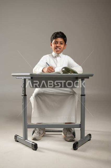 النظر نحو الكاميرا بشكل مباشر بإيماءات السرور, أهمية العلم والتعلم في المجتمع السعودي, بورتريه لطفل عربي خليجي سعودي يرتدي الثوب الأبيض التقليدي يجلس في مقعده ويتابع دروسه, الجد والاجتهاد والمثابرة, خلفية رمادية, محتوى حساس, استخدام حساس