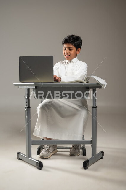 تقدم المملكة العربية السعودية في المجال التعليمي, دمج التكنولوجيا بالتعليم,  بورتريه لطفل عربي خليجي سعودي يرتدي الثوب الأبيض التقليدي يجلس في مقعده ويتابع دروسه عبر الحاسوب المحمول, استخدام تقنيات حديثة في التدريس, خلفية رمادية, محتوى حساس, استخدام حساس