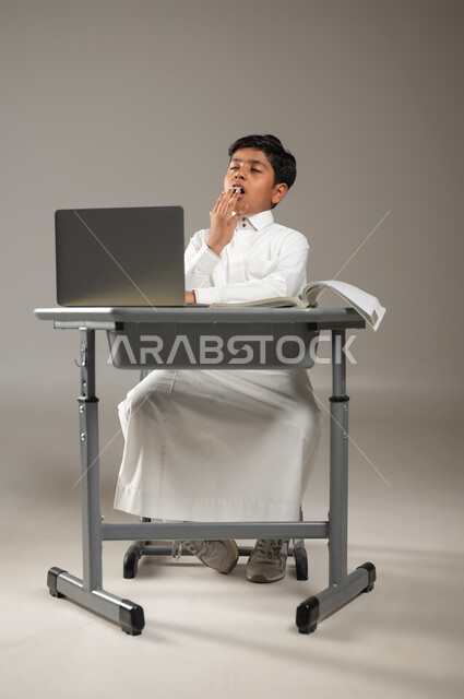 متابعة الدروس بجد واجتهاد, التعلم باستخدام التكنولوجيا, بورتريه لطفل عربي خليجي سعودي يرتدي الثوب الأبيض التقليدي يجلس في مقعده ويتثاءب واضعاً يده على فمه بإيماءات تدل على الشعور بالنعاس, التعب والحاجة إلى النوم و الراحة, خلفية رمادية, محتوى حساس, استخدام حساس