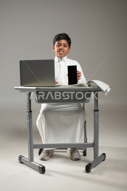 عرض شاشة سوداء للهاتف المحمول, دمج التكنولوجيا بالتعليم في مدارس المملكة العربية السعودية, بورتريه لطفل عربي خليجي سعودي يرتدي الثوب الأبيض التقليدي يجلس في مقعده وأمامه الحاسوب المحمول, النظر إلى الكاميرا بتعبيرات السرور, خلفية رمادية, محتوى حساس, استخدام حساس