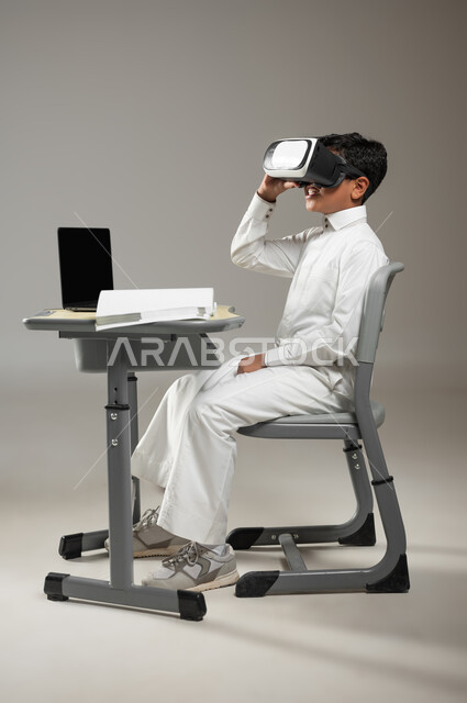 تطور طرائق التعليم في المملكة العربية السعودية, دمج التكنولوجيا  والتقنيات الحديثة بالتعليم, بورتريه لطفل عربي خليجي سعودي يرتدي الثوب الأبيض التقليدي يجلس في مقعده ويلبس نظارة العالم الافتراضي ثلاثية الأبعاد, خلفية رمادية, محتوى حساس, استخدام حساس