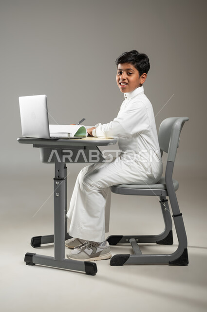 أهمية العلم والتعلم في المملكة العربية السعودبة, دمج التكنولوجيا بالتعليم,, بورتريه لطفل عربي خليجي سعودي يرتدي الثوب الأبيض التقليدي يجلس في مقعده ويتابع دروسه عبر الحاسوب المحمول, النظر إلى الكاميرا بإيماءات السرور, خلفية رمادية, محتوى حساس, استخدام حساس