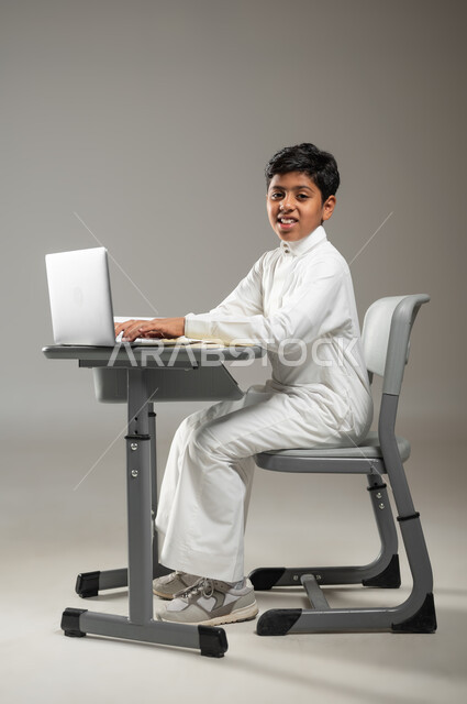 النظر إلى الكاميرا بإيماءات السرور, التعلم باستخدام التقنيات الحديثة عبر الحاسوب المحمول, بورتريه لطفل عربي خليجي سعودي يرتدي الثوب الأبيض التقليدي يجلس في مقعده ويتابع دروسه, دمج التكنولوجيا بالتعليم, خلفية رمادية, محتوى حساس, استخدام حساس