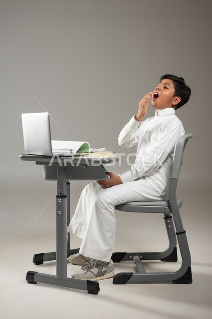 إيماءات تدل على الشعور بالنعاس بعد مذاكرة متعبة, دمج التكنولوجيا بالتعليم, استخدام الحاسوب المحمول في التعلم, بورتريه لطفل عربي خليجي سعودي يرتدي الثوب الأبيض التقليدي يجلس في مقعده ويتثاءب, دروس صعبة وممتعة, خلفية رمادية, محتوى حساس, استخدام حساس