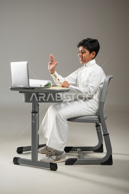 دمج التكنولوجيا بالتعليم, أهمية العلم والتعلم في المملكة العربية السعودية, بورتريه لطفل عربي خليجي سعودي يرتدي الثوب الأبيض التقليدي يجلس في مقعده ويتابع دروسه عبر الحاسوب المحمول, استخدام طرائق حديثة في التدريس, خلفية رمادية, محتوى حساس, استخدام حساس