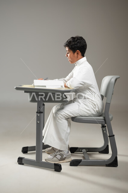 الجد والإجتهاد في الدراسة والمذاكرة من أجل التفوق, أهمية العلم والتعلم, بورتريه لطفل عربي خليجي سعودي يرتدي الثوب الأبيض التقليدي يجلس في مقعده ويقرأ في كتبه, متابعةالمناهج الدراسية بكل مثابرة وعزيمة, خلفية رمادية, محتوى حساس, استخدام حساس