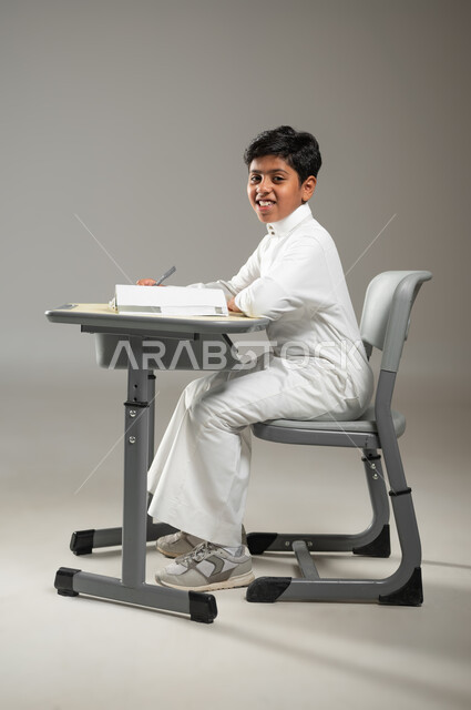 أهمية العلم والتعلم في المجتمع السعودي, الدراسة بجد واجتهاد لتحقيق النجاح, بورتريه لطفل عربي خليجي سعودي يرتدي الثوب الأبيض التقليدي يجلس في مقعده ويتابع دروسه, النظر إلى الكاميرا بإيماءات السعادة, خلفية رمادية, محتوى حساس, استخدام حساس