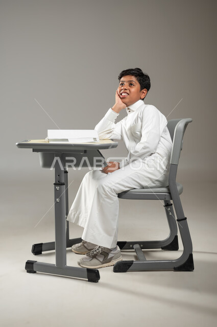 النظر إلى السماء والتحليق في عالم الأحلام المنتظرة, السعي نحو النجاح والتفوق, بورتريه لطفل عربي خليجي سعودي يرتدي الثوب الأبيض التقليدي يجلس في مقعده ويتابع دروسه, النظر إلى الأفق بإيماءات السعادة, أهمية العلم والتعليم, خلفية رمادية, محتوى حساس, استخدام حساس