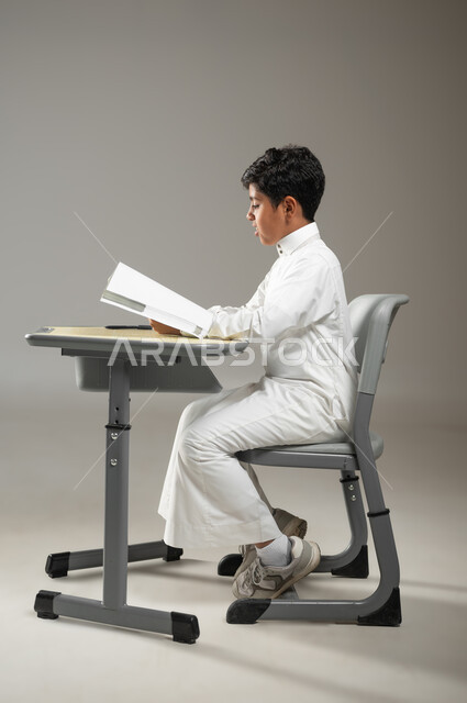 السعي إلى النجاح عن طريق الاجتهاد في الدراسة, أهمية العلم والتعلم, بورتريه لطفل عربي خليجي سعودي يرتدي الثوب الأبيض التقليدي يجلس في مقعده ويتابع دروسه, النظر إلى الأوراق والإمساك بها تعبيراً عن الجد والمثابرة, خلفية رمادية, محتوى حساس, استخدام حساس