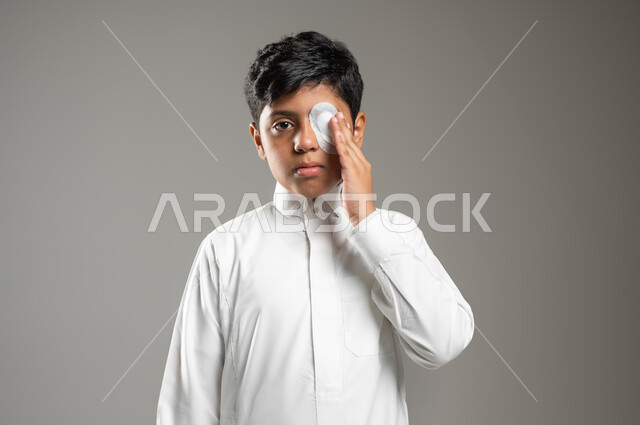 مفهوم الصحة والمرض, النظر إلى الكاميرا بإيماءات الحزن والألم, بورتريه لطفل عربي خليجي سعودي يرتدي الثوب الأبيض التقليدي ويضع يده على عينه المصابة, تغطية العين بضماد طبي من أجل تقوية النظر, التضميد الصحيح للجراح, خلفية رمادية, محتوى حساس, استخدام حساس