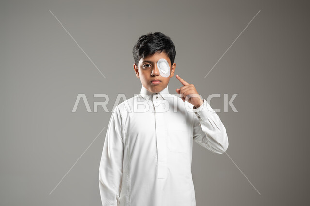 النظر إلى الكاميرا والإشارة إلى العين بإيماءات الحزن, بورتريه لطفل عربي خليجي سعودي يرتدي الثوب الأبيض التقليدي, تغطية العين بضماد طبي من أجل تقوية النظر, التضميد الصحيح للجراح, خلفية رمادية, محتوى حساس, استخدام حساس