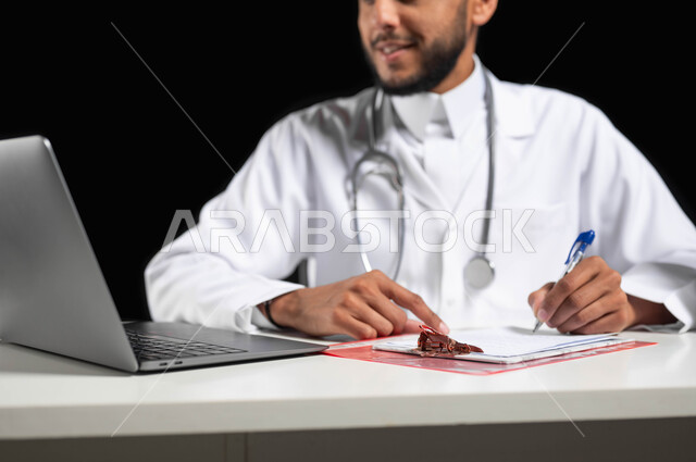 دمج العمل مع التكنولوجيا, تقديم الخدمات الطبية للمرضى و متابعة حالاتهم عبر الحاسوب المحمول, بورتريه لطبيب عربي خليجي سعودي مبتسم يرتدي المعطف و السماعة الطبية يقوم بكتابة وتدوين  التقارير والحالات الصحيه، خلفية سوداء, محتوى حساس, استخدام حساس