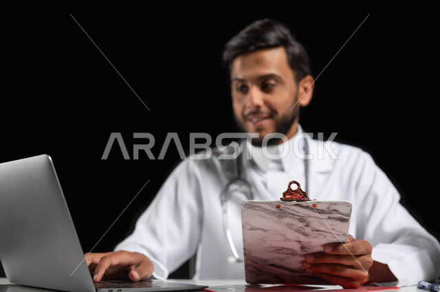 تقديم الخدمات الطبية للمرضى, بورتريه مقرب لطبيب عربي خليجي سعودي يرتدي المعطف الأبيض يقوم بقراءة  التقارير والحالات الصحيه,  استعمال الحاسوب المحمول في المجال الطبي، استخدام حقيبة الاسعافات الاوليه، خلفية سوداء, محتوى حساس, استخدام حساس