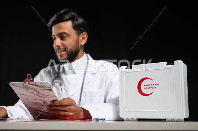 العمل في القطاع الصحي و تقديم الخدمات للمرضى, بورتريه مقرب لطبيب عربي خليجي سعودي يرتدي المعطف الأبيض يقوم بقراءة  التقارير والحالات الصحيه,  استخدام الحاسوب المحمول في المجال الطبي، استخدام حقيبة الاسعافات الاوليه، خلفية سوداء, محتوى حساس, استخدام حساس