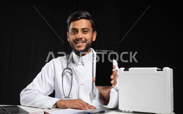 استخدام حقيبة الاسعافات الاوليه والتكنولوجيا في المجال الطبي، العمل في القطاع الصحي، بورتريه مقرب لطبيب عربي خليجي سعودي يرتدي المعطف الطبي يقوم بعرض شاشة سوداء فارغه عبر الهاتف المحمول، خلفية سوداء، استخدام حساس, محتوى حساس