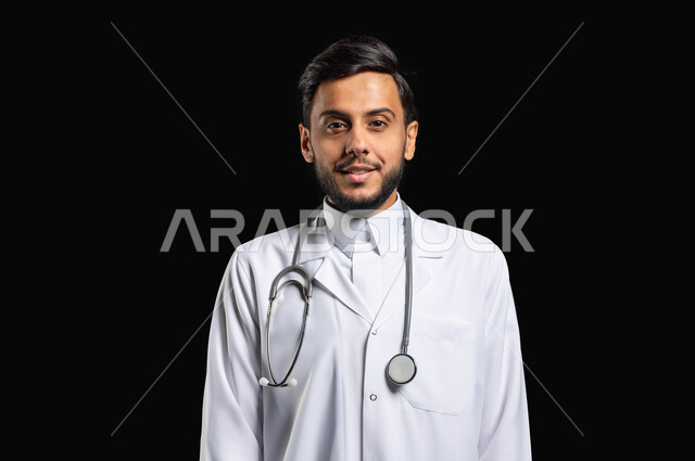 النظر الى الكاميرا بايماءات البشاشه، العمل في القطاع الطبي، بورتريه مقرب لطبيب عربي خليجي سعودي مبتسم يرتدي المعطف مع السماعه الطبيه يقف باستقامة، خلفية سوداء, محتوى حساس, استخدام حساس