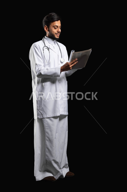 ايماءات البشاشه اثناء العمل في القطاع الطبي، بورتريه طبيب عربي خليجي سعودي مبتسم يرتدي المعطف و السماعة الطبية يقوم بقراءة التقارير الطبية، صورة الجسم بالطول الكامل، خلفية سوداء, محتوى حساس, استخدام حساس