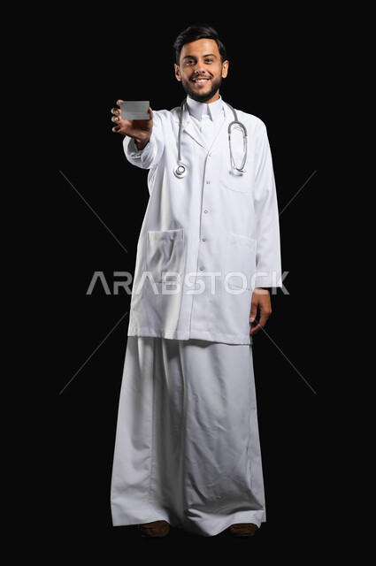 الوقوف باستقامة مع النظر الى الكاميرا بتعابير الرضا والسرور، الرعايه الصحيه في المملكة العربية السعوديه، بورتريه لطبيب عربي خليجي سعودي مبتسم يرتدي الثوب التقليدي مع المعطف الطبي يعرض البطاقة الشخصية لتعريف عن نفسه، صورة الجسم بالطول الكامل، خلفية سوداء، محتوى حساس، استخدام حساس