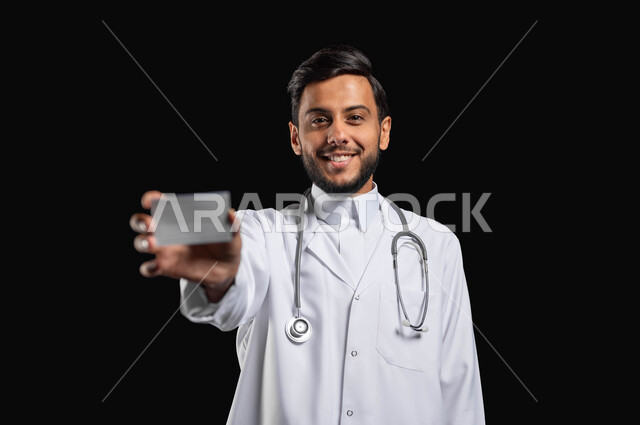 مفهوم الطب و الرعاية الصحية، النظر الى الكاميرا بتعابير السرور، بورتريه مقرب لطبيب عربي خليجي سعودي يقوم بعرض بطاقة شخصية بيضاء، خلفية سوداء، محتوى حساس، استخدام حساس