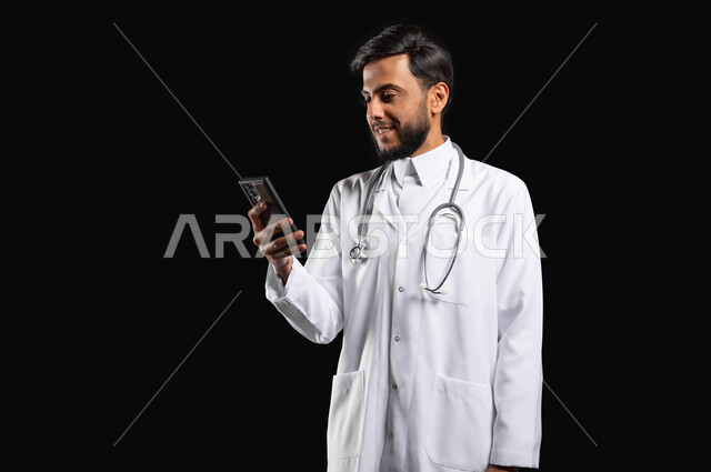 The concept of medicine and health care, work in the health sector, the development of medicine through technology, portrait of a Saudi Gulf Arab doctor wearing a coat with a stethoscope using a mobile phone, black background, sensitive content, sensitive use