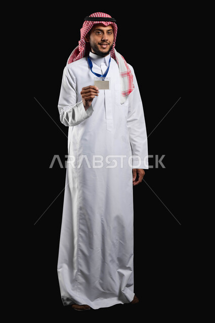 الوقوف باستقامة مع النظر الى الكاميرا بتعابير الرضا والسرور، بورتريه لشاب عربي خليجي سعودي مبتسم يرتدي الزي التقليدي والشماغ يضع البطاقة الشخصية لتعريف عن نفسه، صورة الجسم بالطول الكامل، خلفية سوداء، محتوى حساس، استخدام حساس