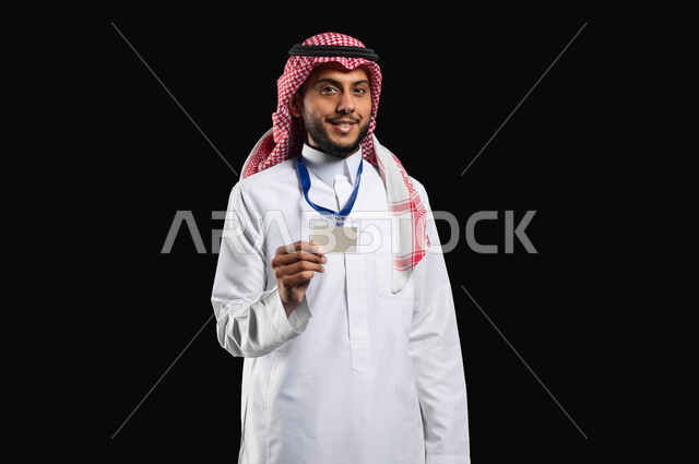 مفهوم الاناقة والاهتمام بالمظهر الخارجي، الوقوف بثقة واستقامة، أهمية التعريف عن النفس، بورتريه لشاب عربي خليجي سعودي مبتسم يرتدي الزي التقليدي والشماغ يحمل البطاقة الشخصية بايماءات الرضا والسرور، خلفية سوداء، محتوى حساس، استخدام حساس