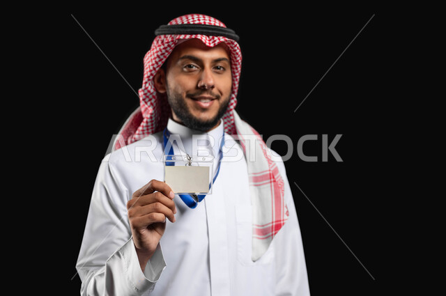 اظهار البطاقة الشخصية،مفهوم الاناقة والاهتمام بالمظهر الخارجي، الوقوف بثقة واستقامة، بورتريه لشاب عربي خليجي سعودي مبتسم يرتدي الزي التقليدي والشماغ ينظر إلى الكاميرا بايماءات الرضا والسرور، خلفية سوداء، محتوى حساس، استخدام حساس