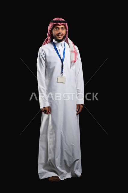 ارتداء الازياء الشعبية التقليدية، الوقوف باستقامة مع النظر الى الكاميرا بتعابير الرضا والسرور، بورتريه لشاب عربي خليجي سعودي مبتسم يرتدي الزي التقليدي والشماغ يضع البطاقة الشخصية لتعريف عن نفسه، صورة الجسم بالطول الكامل، خلفية سوداء، محتوى حساس، استخدام حساس
