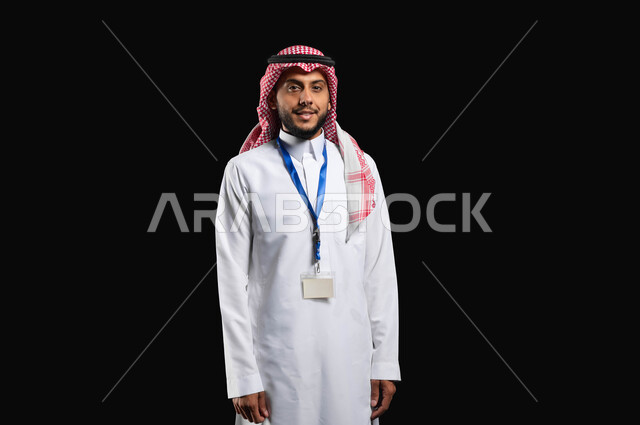 مفهوم الاناقة والاهتمام بالمظهر الخارجي، الوقوف بثقة واستقامة، أهمية البطاقة الشخصية لتعرف على الشخص، بورتريه لشاب عربي خليجي سعودي مبتسم يرتدي الزي التقليدي والشماغ ينظر إلى الكاميرا بايماءات الرضا والسرور، خلفية سوداء، محتوى حساس، استخدام حساس