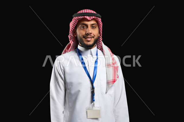 النظر الى الكاميرا بتعابير الفرح والسرور، مفهوم الاناقة والاهتمام بالمظهر الخارجي، أهمية البطاقة الشخصية لتعرف ع الشخص، بورتريه مقرب لشاب عربي خليجي سعودي مبتسم يرتدي الزي التقليدي والشماغ يقف باستقامة بايماءات الرضا والثقة، خلفية سوداء، محتوى حساس، استخدام حساس
