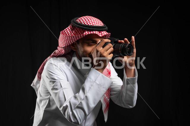 موهبة احتراف فن التصوير, بورتريه مقرب لشاب عربي خليجي سعودي يرتدي الزي التقليدي والشماغ، يحمل بيده كاميرا التصوير عالية الدقة ويلتقط الصور، التصوير الفوتوغرافي، مصورين سعوديين ، كاميرات حديثة ومتطورة, استخدام التقنيات التكنولوجية في مجال التصوير, خلفية سوداء
